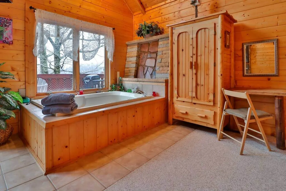 A tranquil bathroom scene showing a bathtub positioned by a window offering scenic natural views. The space includes rustic wooden accents, a stone fireplace, and a vanity table, fostering a sense of comfort and rejuvenation.