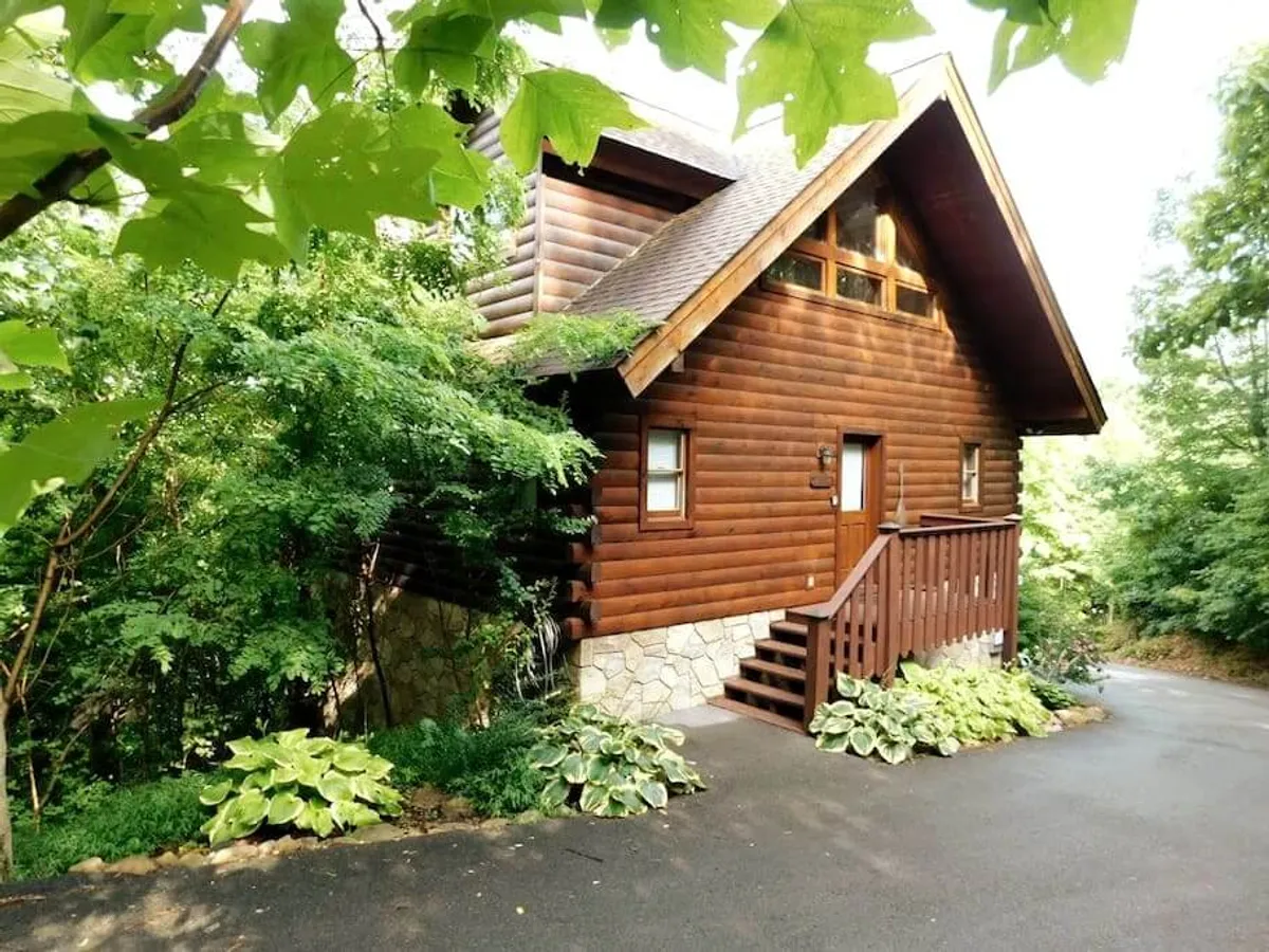 A charming log cabin with a solid stone foundation, surrounded by landscaped gardens and mature trees. A wooden staircase leads up to a porch, inviting guests to relax and enjoy the tranquil setting. The cabin is nestled in nature, offering a sense of seclusion and privacy.