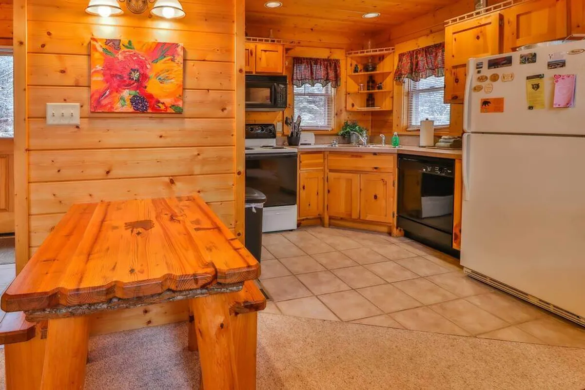 This image highlights a welcoming kitchen and dining area, featuring a sturdy wooden dining table, colorful wall art, kitchen appliances, and wooden cabinetry. Abundant natural light filters into the space, creating an inviting ambiance for shared meals or morning coffee.