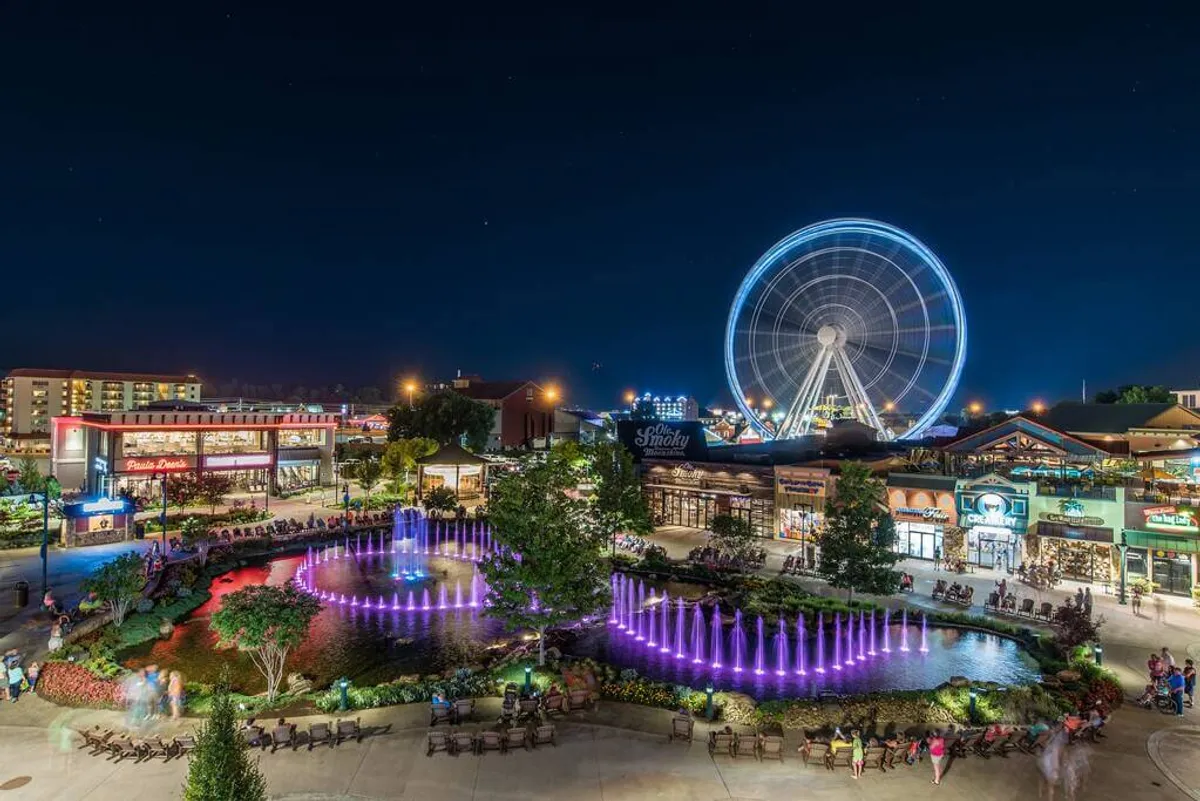 A lively, colorful scene featuring a large Ferris wheel, a playful water fountain, and people enjoying rides, shopping, and outdoor dining. The vibrant atmosphere captures the excitement of exploring local attractions.