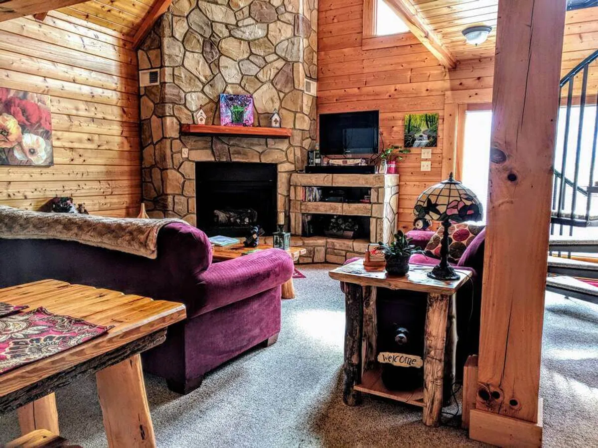 This image presents a living area with a stone fireplace, a richly colored purple sofa, rustic wooden tables, and a decorative lamp, all bathed in warm, welcoming light. The ambiance is both charming and intimate.