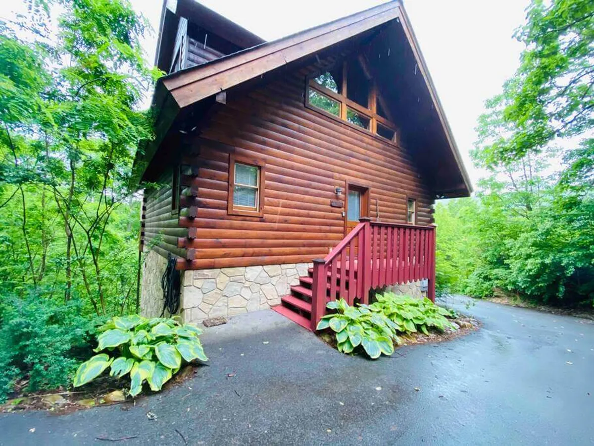 A serene view of the log cabin’s exterior surrounded by lush greenery, featuring a distinctive red staircase and a stone foundation. The spacious porch and tranquil environment hint at the cabin’s privacy, outdoor relaxation, and the peaceful retreat awaiting after days spent exploring.