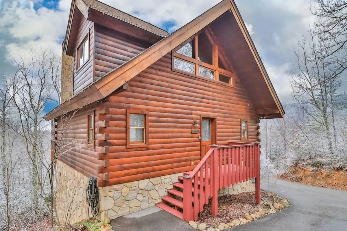 The hero image shows a charming log cabin with inviting red stairs, a sturdy stone foundation, and large windows nestled among the lush woods of Gatlinburg. The scene feels warm and welcoming, suggesting a peaceful retreat in nature.