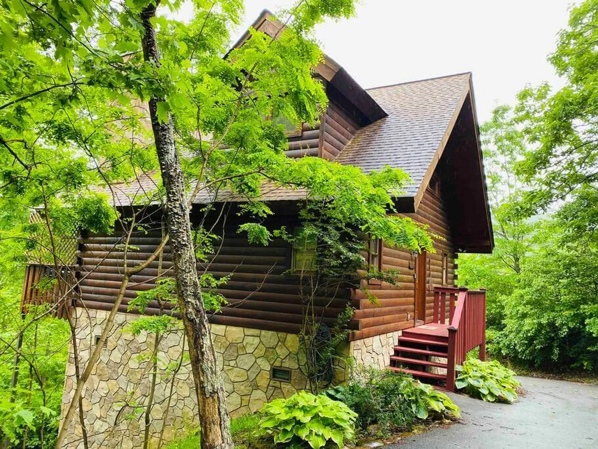A charming log cabin with a stone foundation, wooden steps, and lush greenery around the porch. The exterior evokes a welcoming and cozy atmosphere, ideal for returning to after a day out.