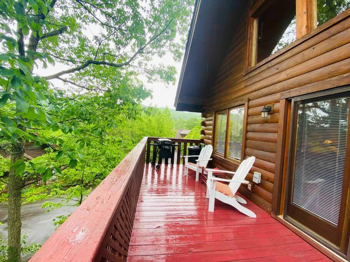 A spacious red wooden deck featuring white Adirondack chairs and an outdoor dining set, all surrounded by lush green foliage. The setting provides a tranquil space to relax and dine while taking in the natural beauty of the outdoors.