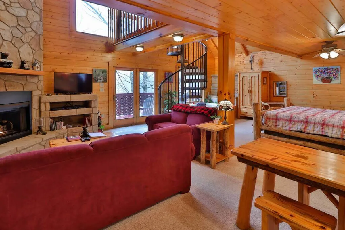 This interior image captures a spacious living room with a stone fireplace, plush sofas, spiral staircase, and a wooden dining table. The setting exudes warmth, cleanliness, and tasteful decor, with a layout ideal for intimate moments and relaxed gatherings.
