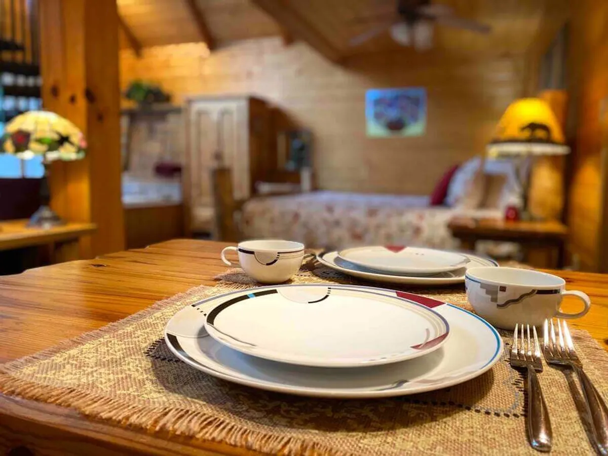 A cozy dining table is thoughtfully set for two, with a decorative lamp casting a gentle glow and a comfortable bed visible in the background. The arrangement underscores intimacy and versatility, ideal for both special breakfasts and romantic dinners.